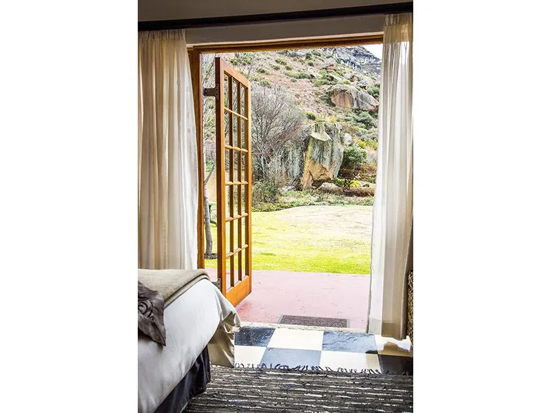 Open door to a green lawn and rocky hillside from a bedroom with curtains