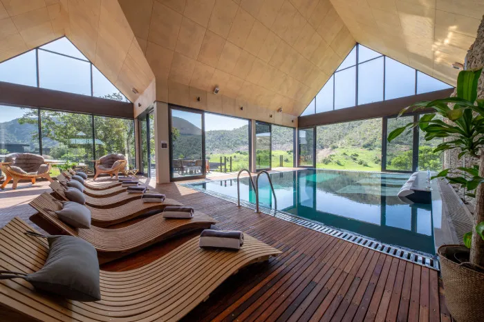 Indoor pool with wooden lounge chairs and large windows overlooking green hills