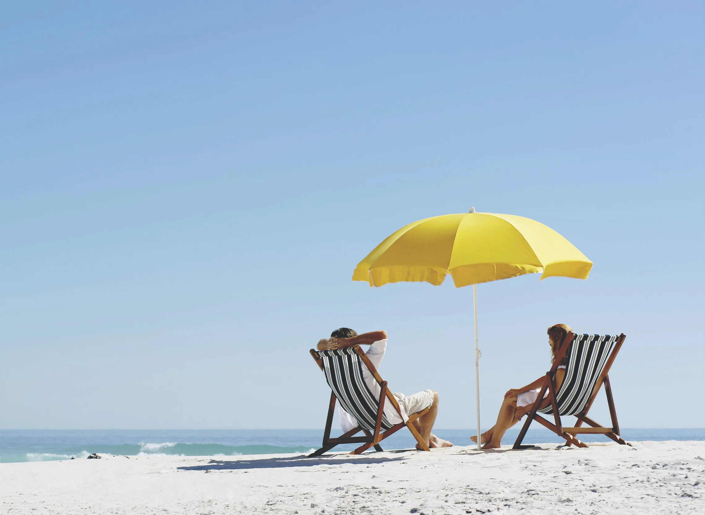 Two people relaxing on beach chairs under a yellow umbrella at the beach