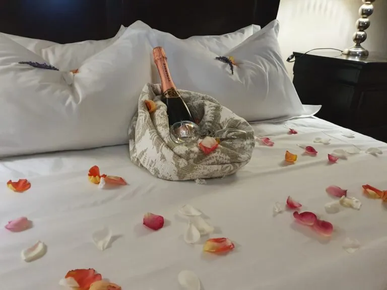 A bottle of champagne in a floral bag on a bed with rose petals