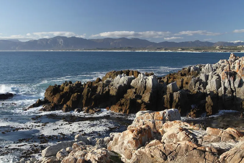 Rocky coastline with ocean waves and distant mountains under a clear sky
