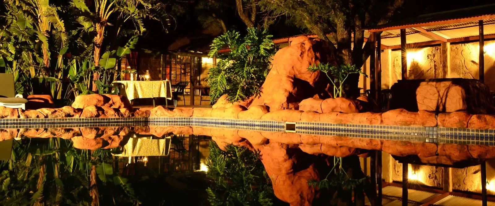 Night view of a pool with rock formations and tropical plants in the background