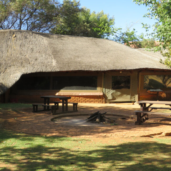 Thatched-roof hut with picnic tables and a fire pit in a garden.