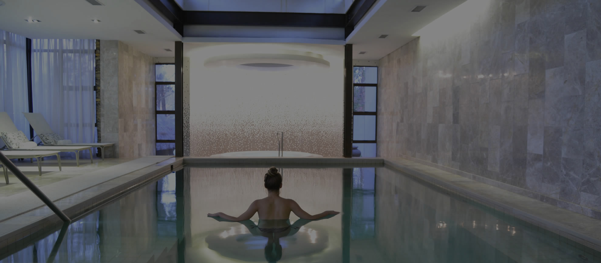 Woman relaxing in an indoor pool with stone walls and large windows.