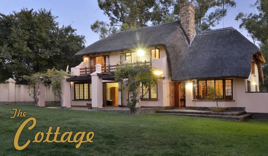 Thatched cottage with lit windows at dusk surrounded by greenery