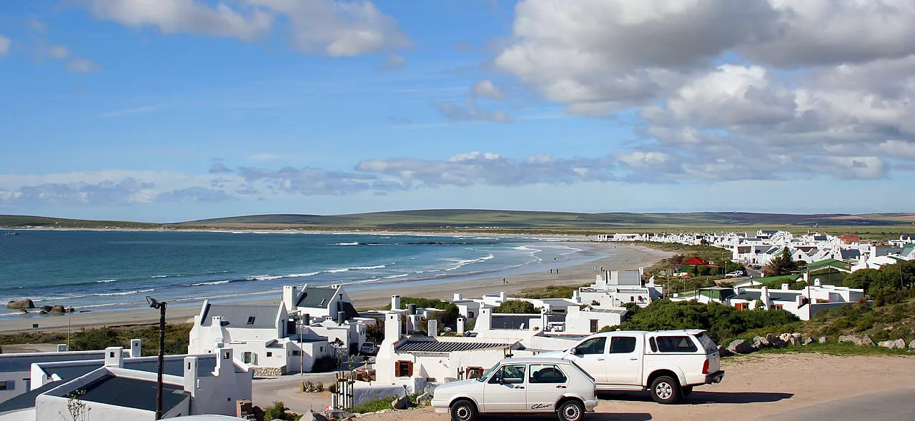 Coastal village with white buildings and parked cars overlooking a sandy beach and ocean