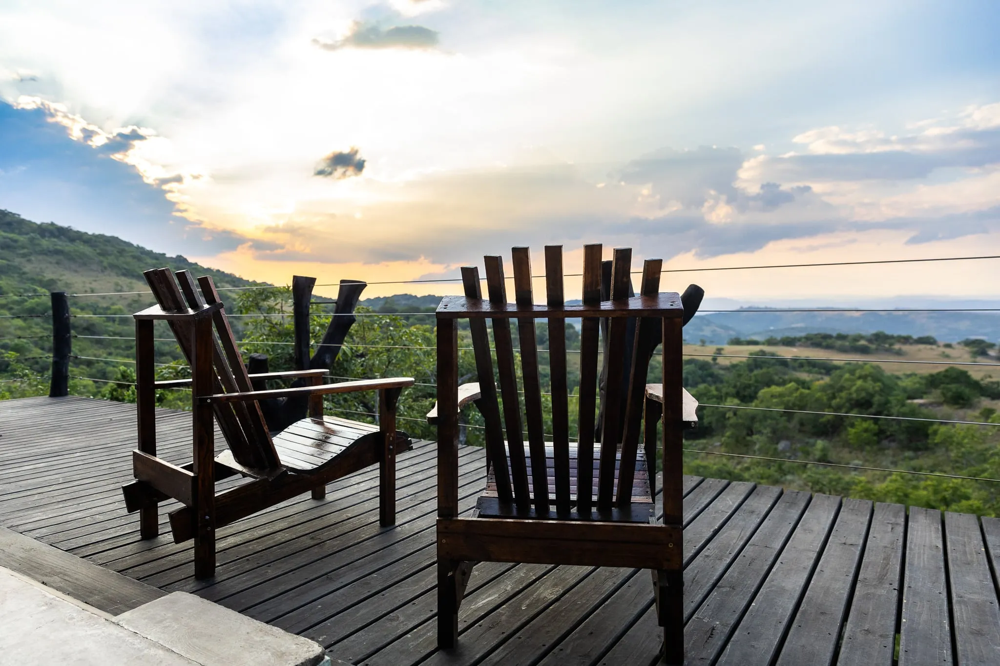 Two wooden chairs on a wooden deck overlooking a scenic landscape at sunset