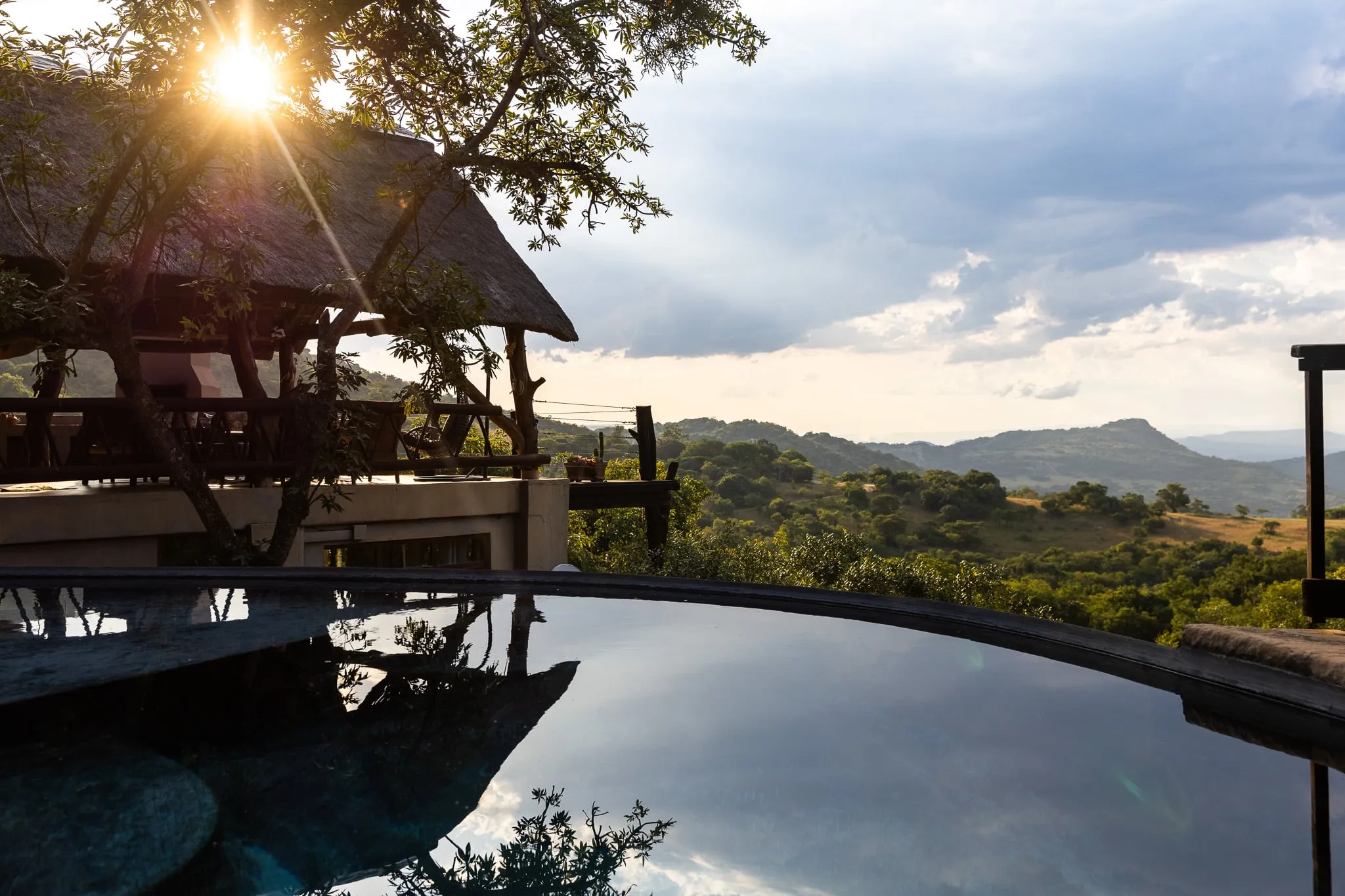 Sunset view over a pool with a thatchedroof structure and distant hills