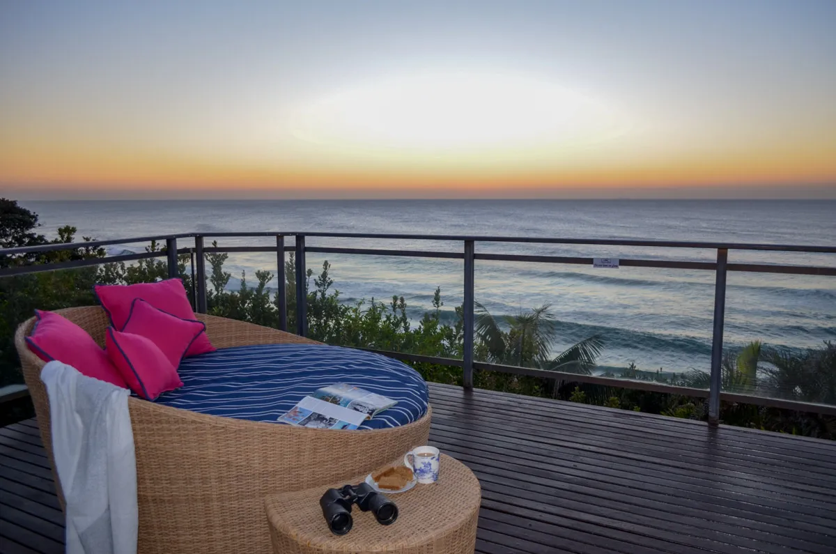 Wicker daybed with pillows on a wooden deck overlooking the ocean at sunset