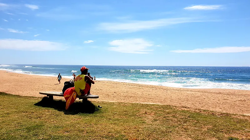Person sitting on a bench overlooking a sandy beach and ocean under a blue sky