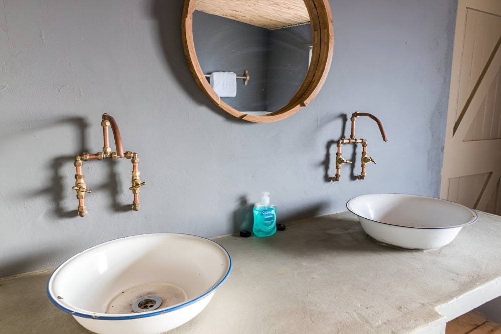 Two white sinks with copper faucets and a round mirror in a rustic bathroom.