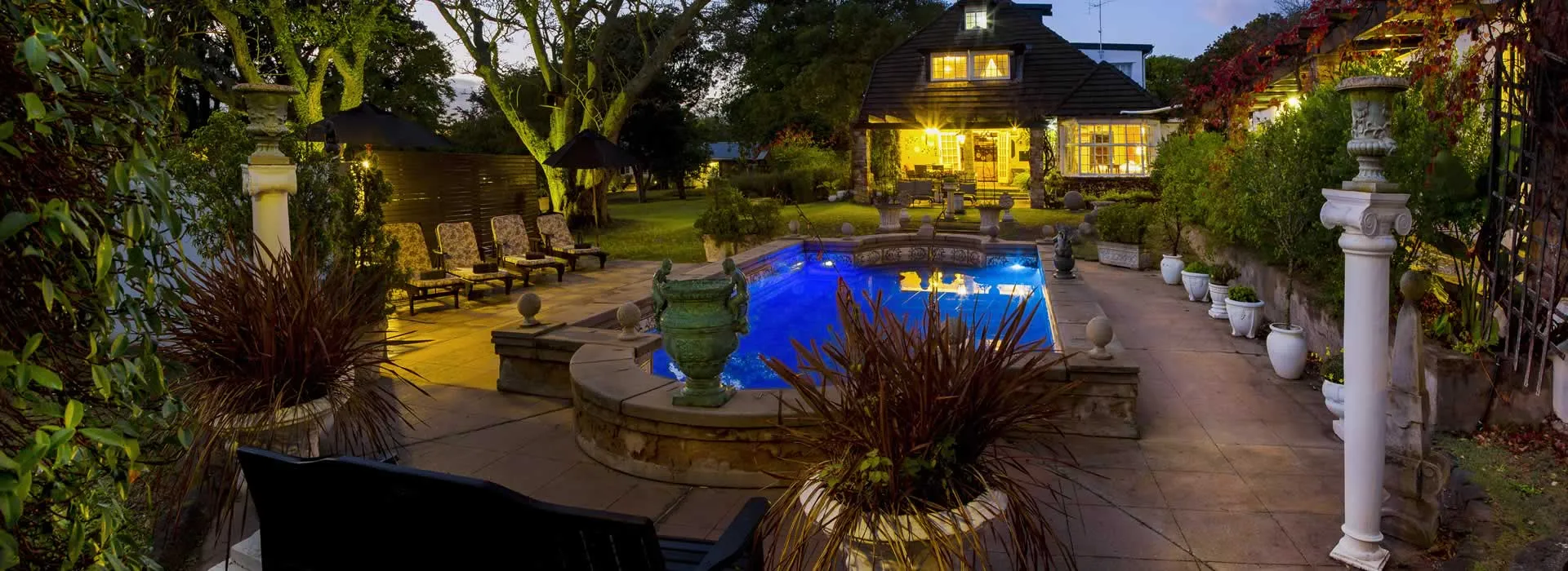 Evening view of a lit pool and house with potted plants and lounge chairs