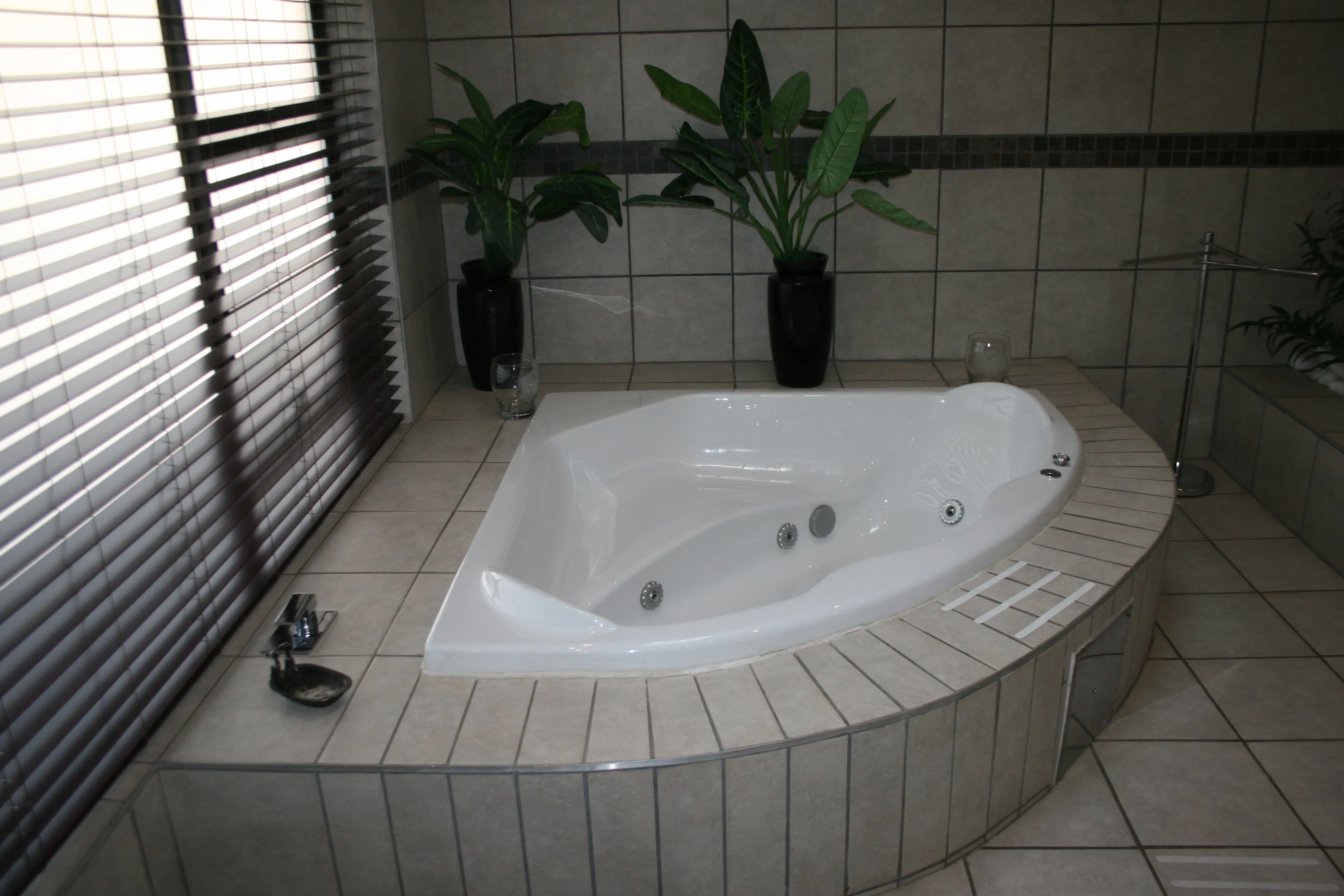 White corner jacuzzi tub in tiled bathroom with plants and window blinds
