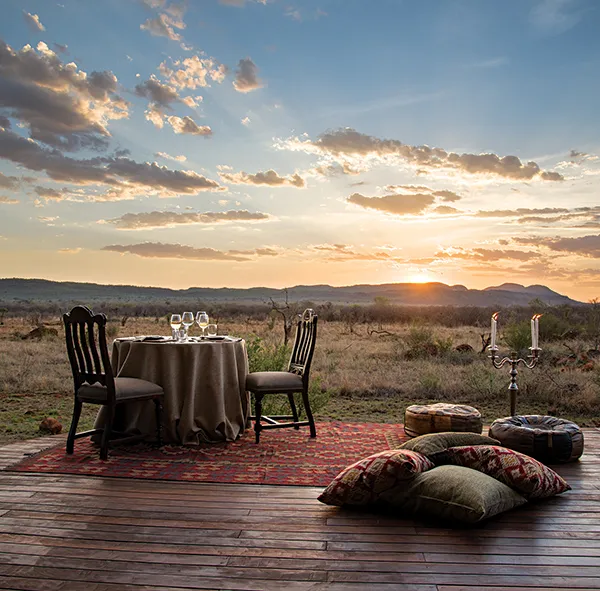Romantic outdoor dinner setup at sunset with table chairs and cushions on a rug