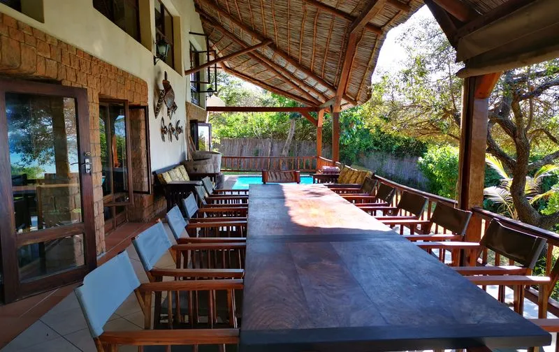 Long wooden table with chairs on a covered patio overlooking a pool and garden