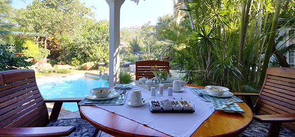 Wooden chairs around a table with breakfast items overlooking a pool and garden