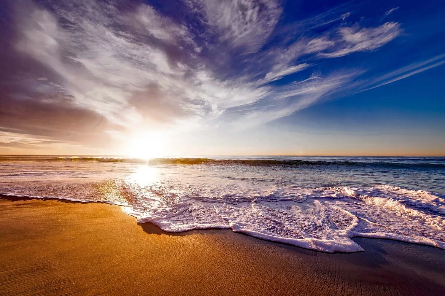 Sunset over ocean with waves crashing onto sandy beach under partly cloudy sky