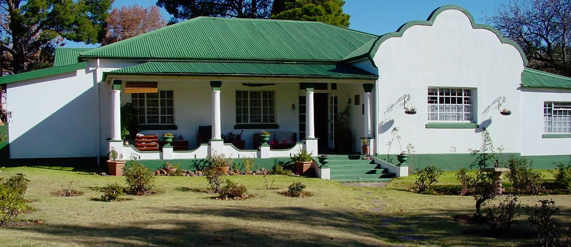Singlestory white house with green roof and veranda surrounded by a garden