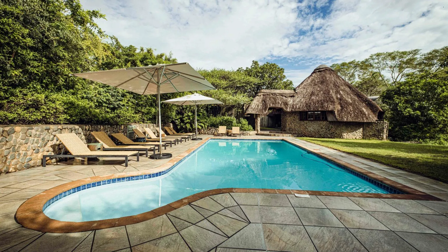 Swimming pool with lounge chairs and umbrellas near a thatchedroof building in a garden