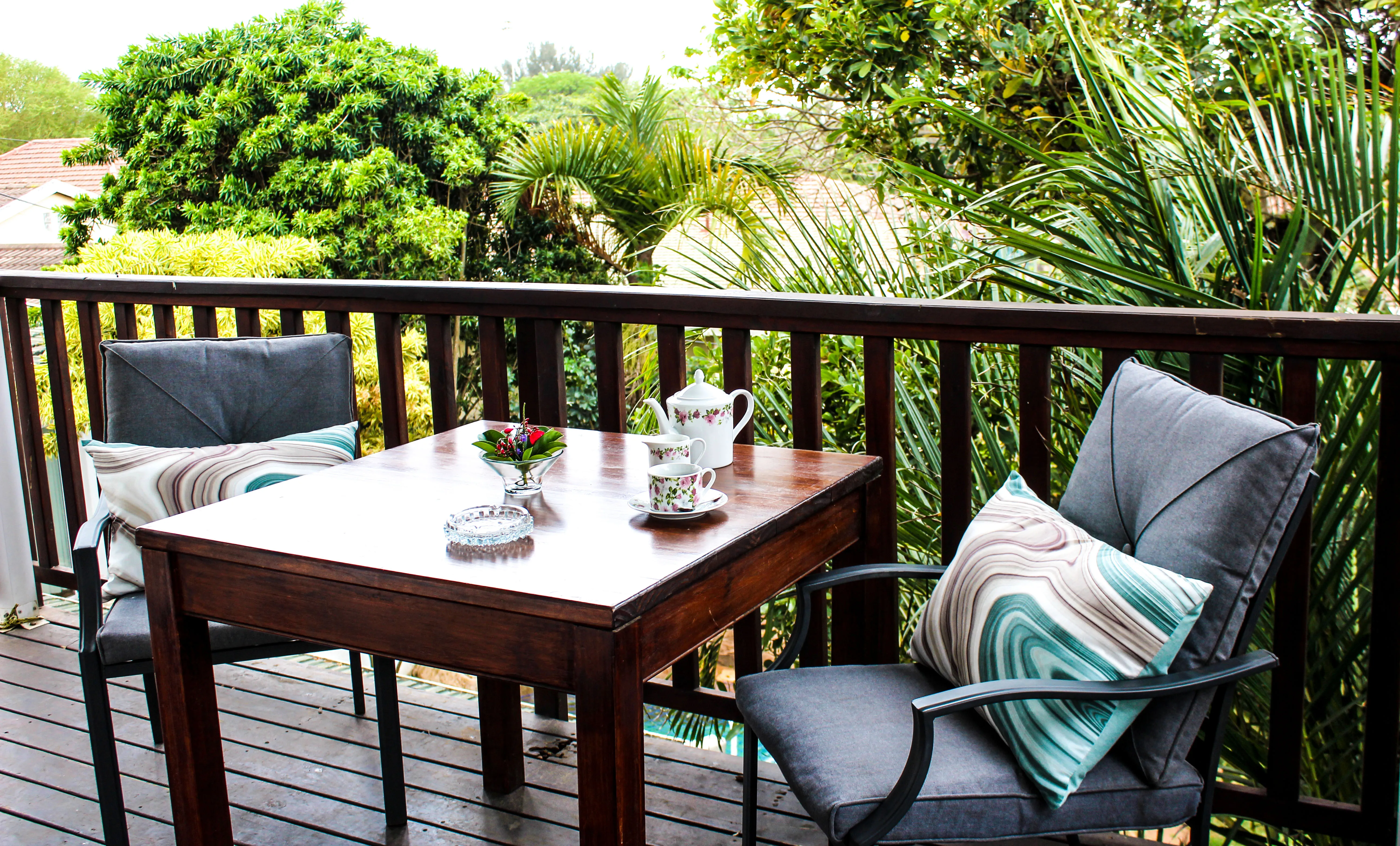 Wooden table with tea set on a deck with two cushioned chairs and greenery