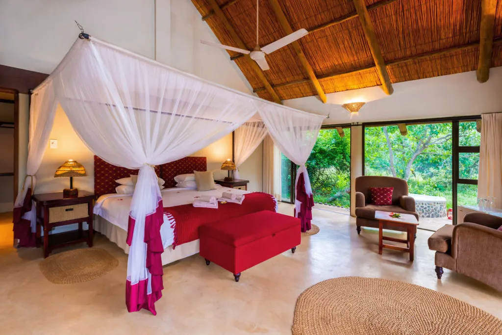 Bedroom with canopy bed red ottoman and large windows overlooking greenery