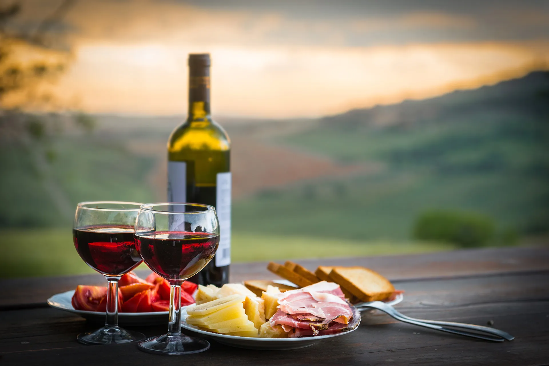 Two glasses of red wine cheese and meat on a table with a scenic background