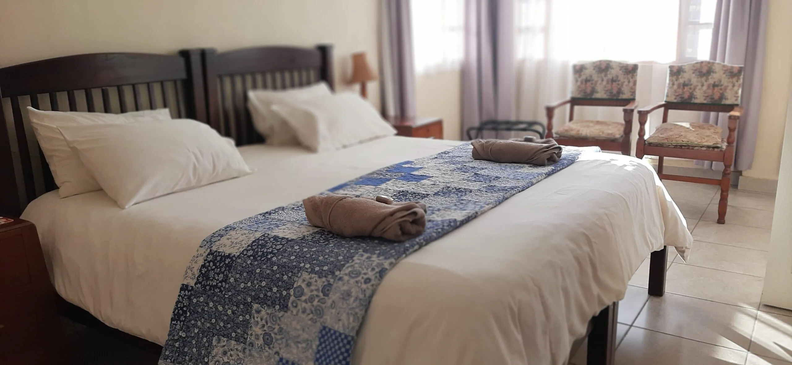 A neatly made bed in a bright bedroom with two chairs and curtains