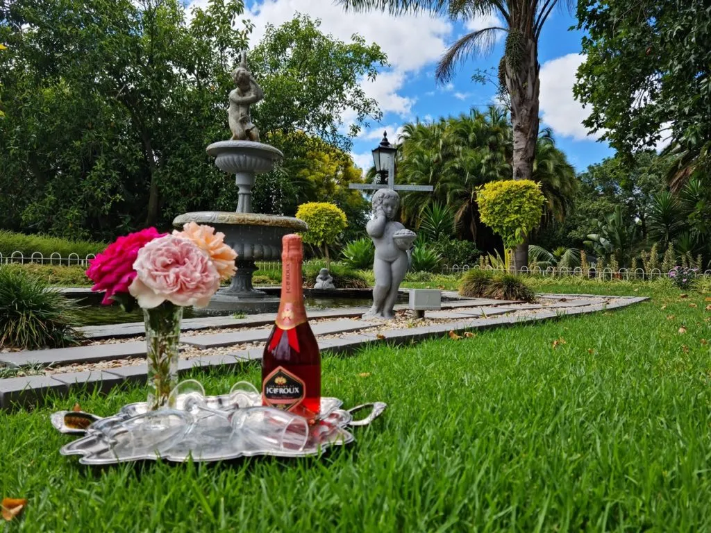 A bottle of wine and flowers on a tray in a garden with statues