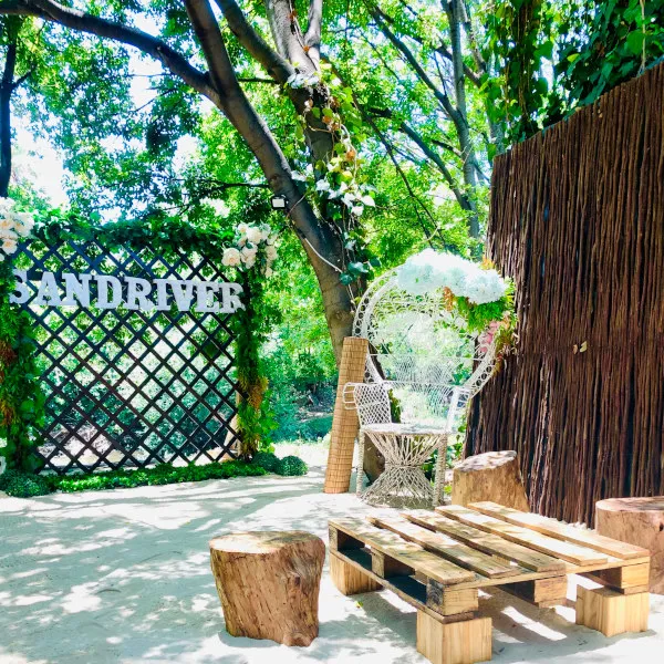 Wooden sign SANDRIVER in a lush garden with wicker chairs and logs