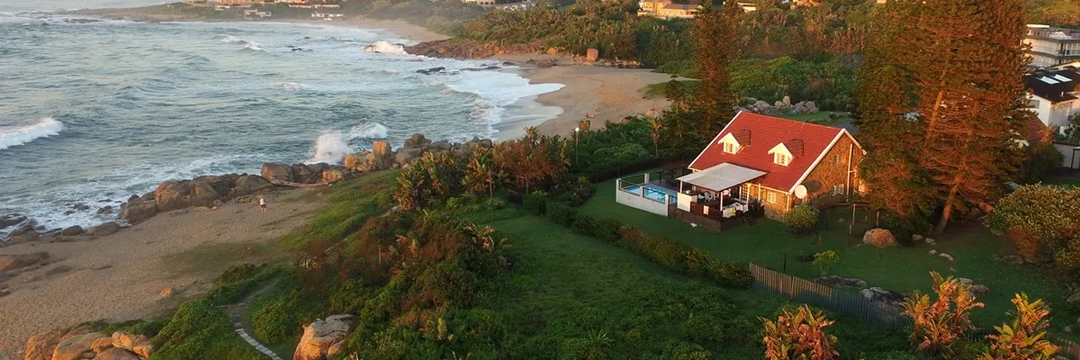 House with red roof near a beach surrounded by greenery and ocean waves