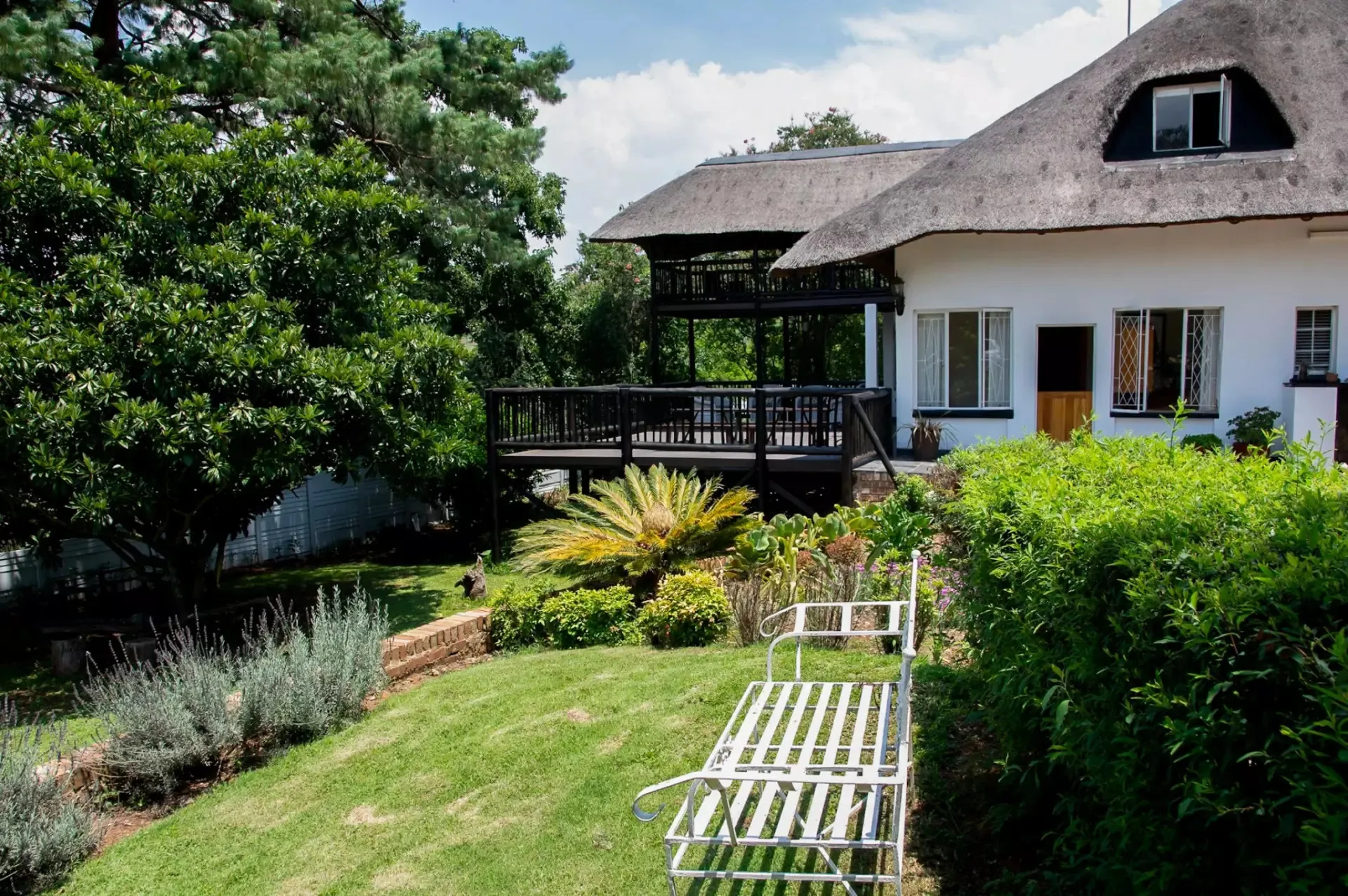 White bench in front of a thatchedroof house with a garden