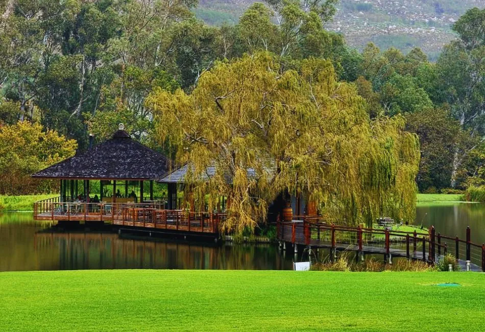 Wooden gazebo by a lake with a bridge surrounded by trees and greenery