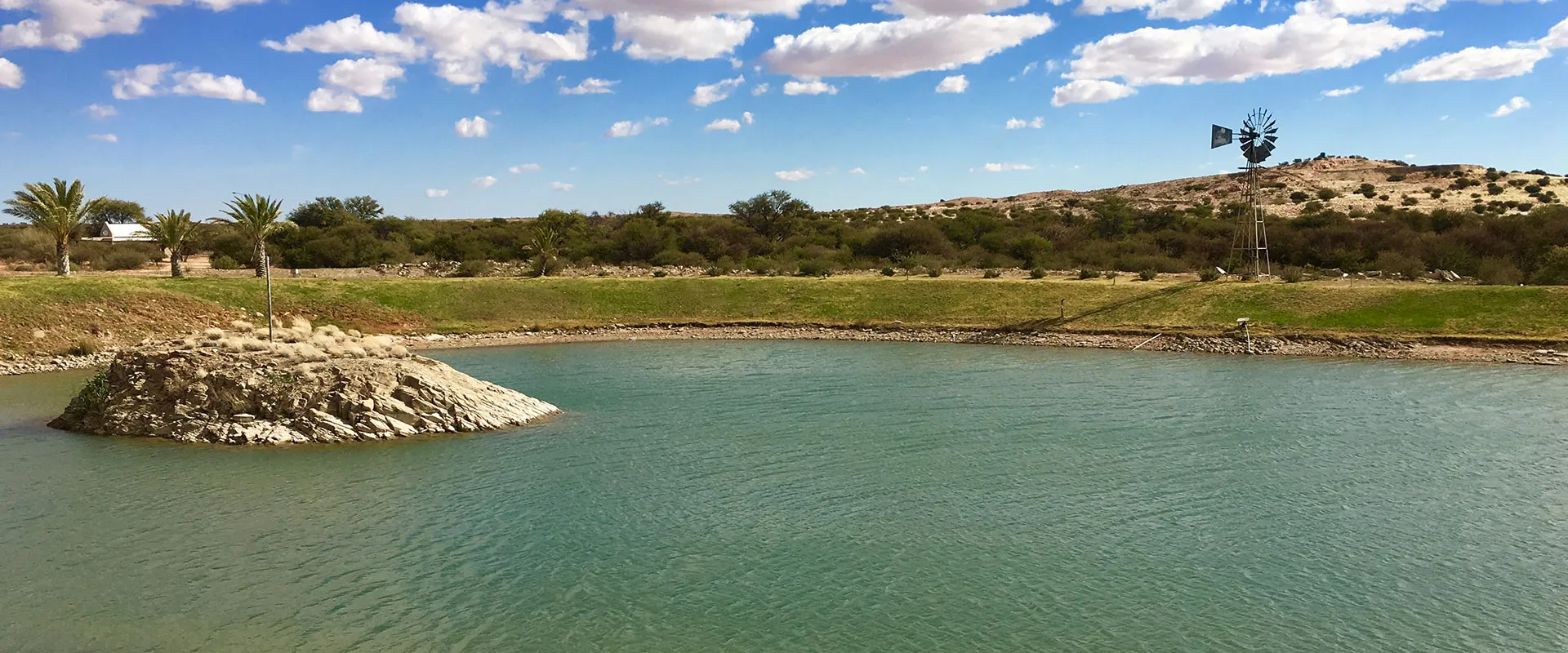 A pond with a small rocky island windmill and palm trees in a hilly landscape