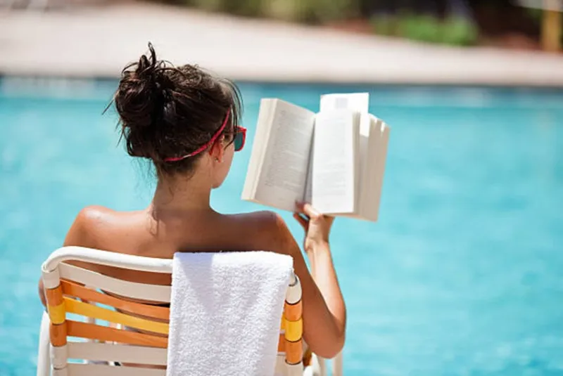 Woman reading a book by a pool sitting on a lounge chair