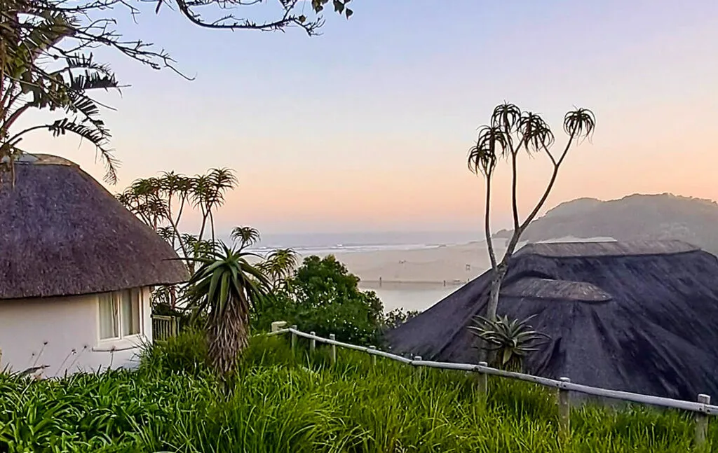 Thatchedroof huts with palm trees overlooking a distant beach at sunset