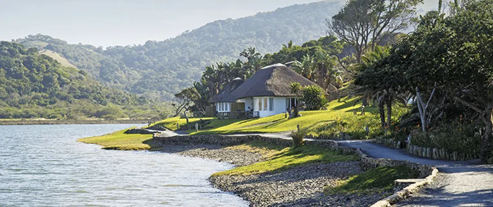 A lakeside cottage with a thatched roof surrounded by greenery and hills