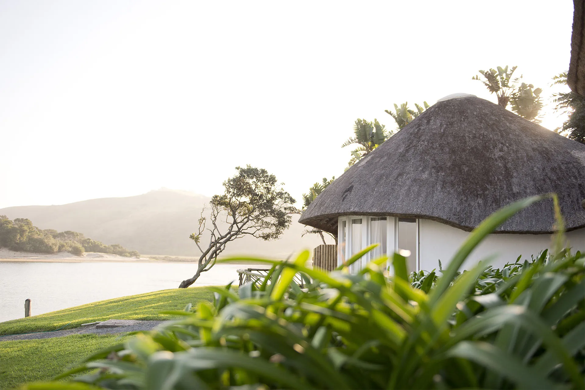 Thatchedroof hut with greenery in front overlooking a lake and hills
