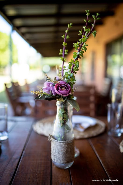 Purple flowers in a glass bottle on a wooden table outdoors.