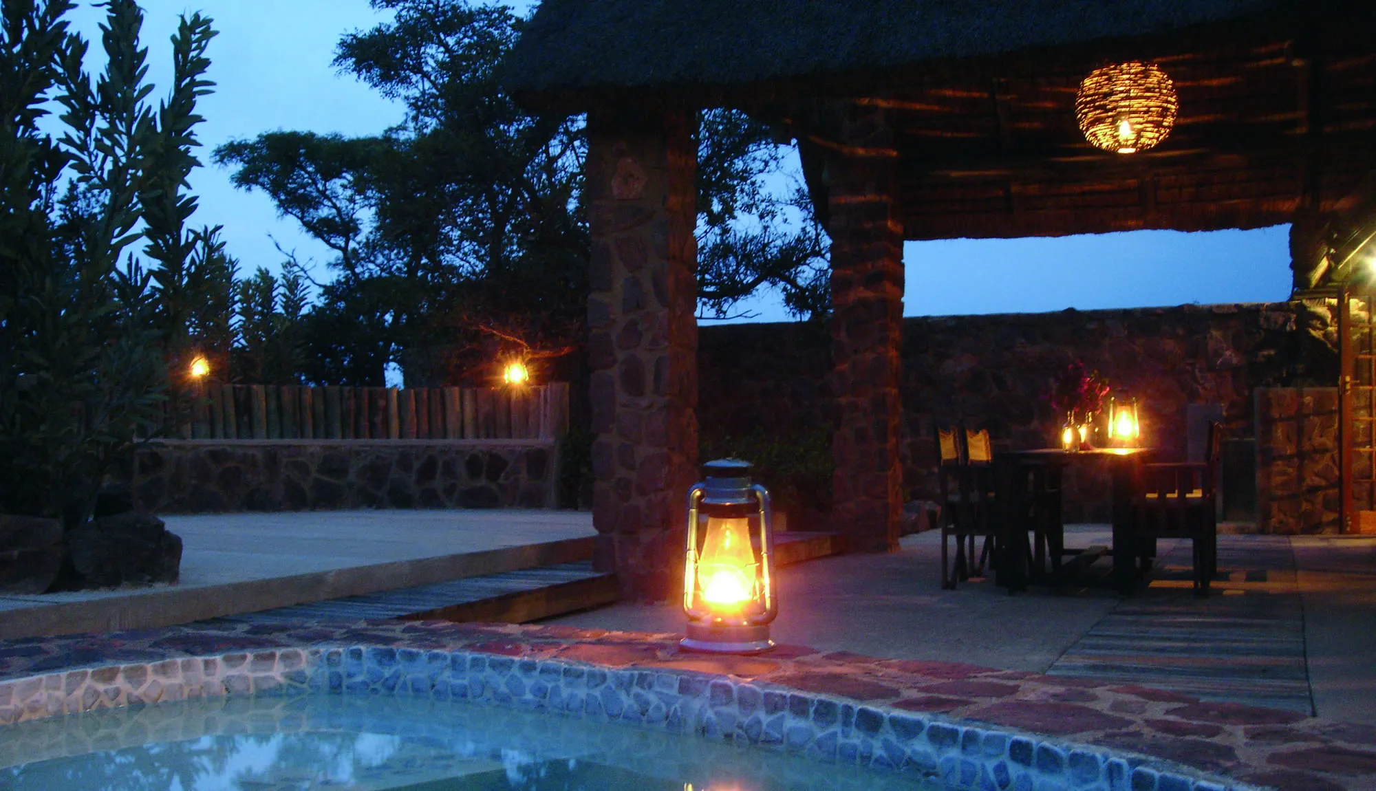 Evening scene with lit lantern near pool and outdoor dining area under thatched roof