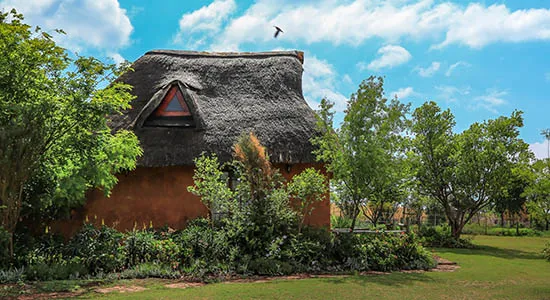 Traditional thatchedroof hut surrounded by greenery under a blue sky with clouds